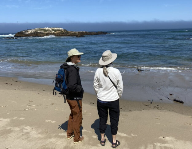 Two middle-aged women in hiking/outdoor gear standing on a beach looking out at the Pacific