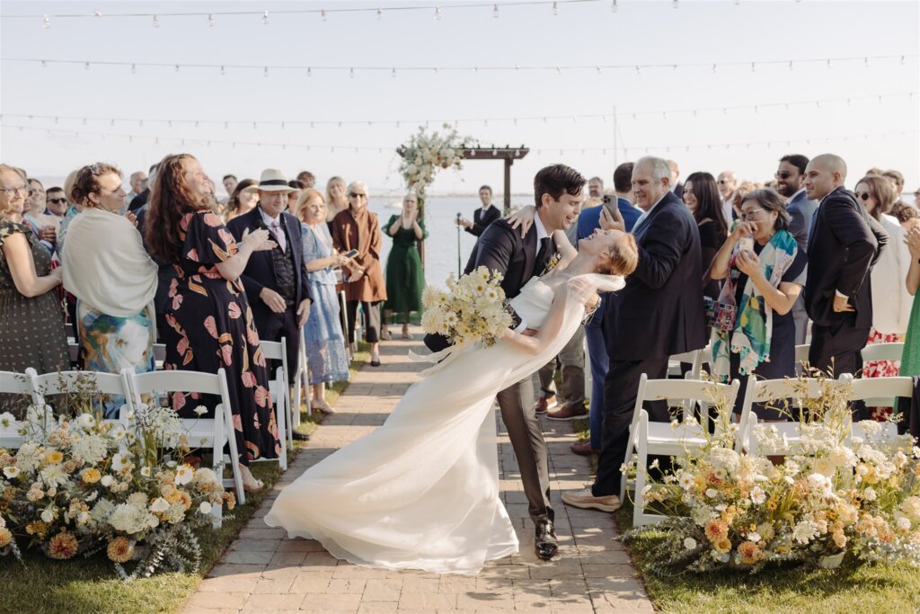 A groom dips his new wife for a kiss, as they exist the aisle and guests stand to clap.