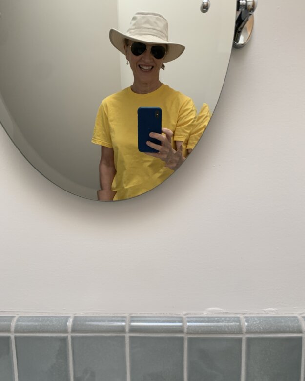 Woman wearing Tilley hat, Ray-Bans and a yellow shirt, looking in mirror 