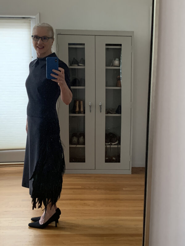An older woman modeling a black silk faille dress with a peacock feather and beads ornament at the hip.